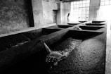 A black and white photograph of an industrial-looking indoor space with large, rectangular concrete basins. Water flows through spouts into the basins, creating ripples on the water's surface. Sunlight filters through large, paned windows on the right, casting light and shadows across the room. A solitary person stands near the windows, adding a human element to the stark environment.