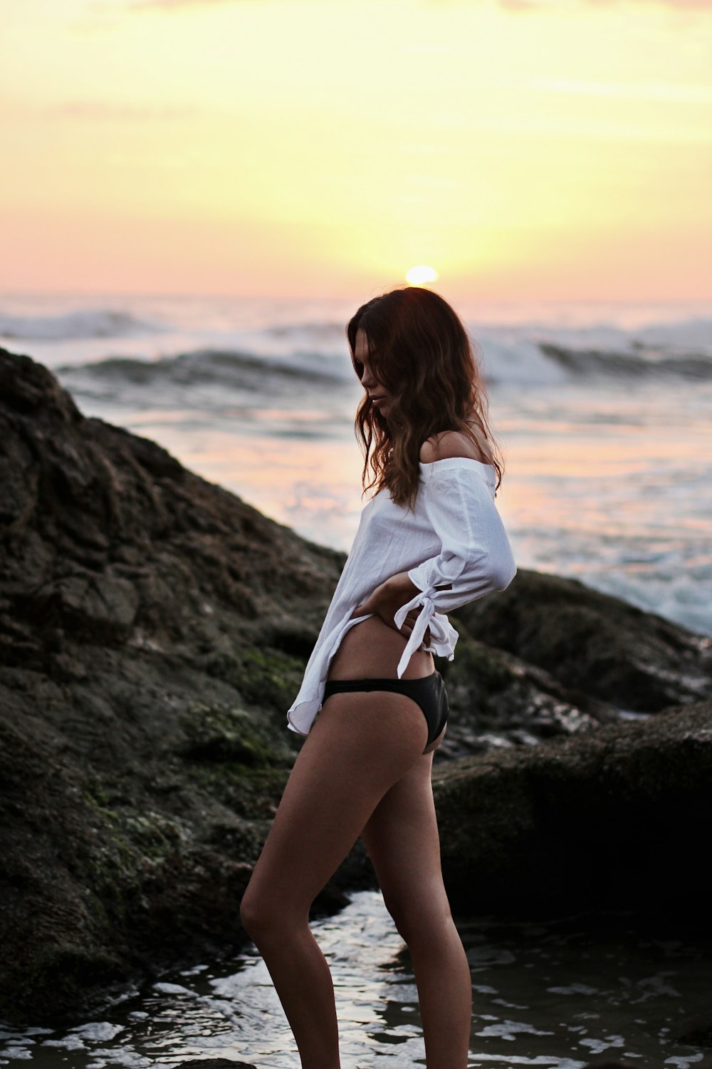 mulher vestindo camisa branca fora dos ombros e calcinha preta em pé na praia com grande rocha durante a hora dourada