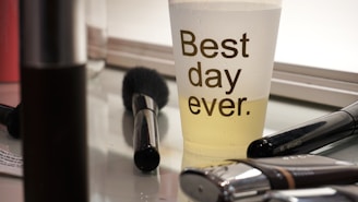 A clear plastic cup with the words 'Best day ever' printed on it is half-filled with a yellow liquid and placed on a reflective surface. Surrounding the cup are several makeup tools including brushes and containers, suggesting a makeup workspace.