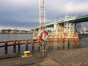A waterfront scene featuring a large bridge with an industrial and modern design spanning across a body of water. In the foreground, there is a lifebuoy attached to a pole, and various construction and utility equipment are visible near the water's edge. The sky is overcast, and the area appears to be in an urban setting with some distant buildings and trees visible.