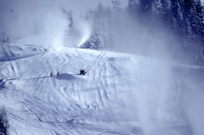 Snowmaking machines operating on a ski slope surrounded by pine trees.