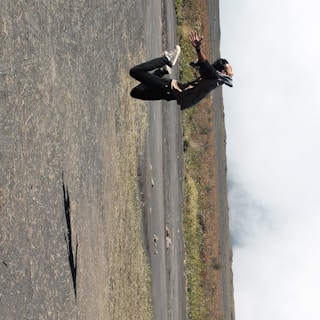 A person is captured in mid-air, performing a high jump with arms outstretched and legs bent. The setting appears to be an open, desolate landscape with a gray, rocky ground and sparse vegetation. The sky is cloudy, and the person's shadow is visible on the ground.