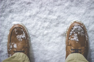 Brown leather shoes dusted with freshly fallen snow, positioned on a snowy ground. The surrounding snow appears clean and untouched, contrasting with the rugged texture of the shoes.