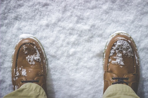 Brown leather shoes dusted with freshly fallen snow, positioned on a snowy ground. The surrounding snow appears clean and untouched, contrasting with the rugged texture of the shoes.