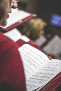 A close-up view of an open music score held by a person dressed in red. Other blurred figures in similar attire are visible in the background, suggesting a choir or group performance. The sheet music is clear and prominently displayed.