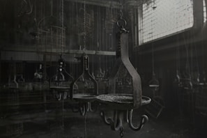 A dimly lit industrial setting with a series of metal hooks and chains hanging from the ceiling, perhaps resembling a workshop or storage area. The room appears abandoned with dust covering the surfaces and low lighting casting shadows across the scene. The atmosphere is eerie and somewhat desolate.