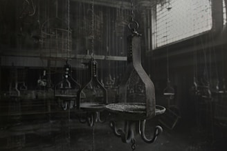 A dimly lit industrial setting with a series of metal hooks and chains hanging from the ceiling, perhaps resembling a workshop or storage area. The room appears abandoned with dust covering the surfaces and low lighting casting shadows across the scene. The atmosphere is eerie and somewhat desolate.