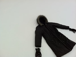 A dark winter coat with fur trim on the hood lies on a flat, snowy surface. The coat appears to have one glove at each sleeve end, suggesting it may have been laid out to mimic a person making a snow angel. The snow is smooth, contrasting with the dark color of the coat.