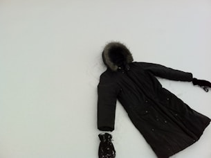 A dark winter coat with fur trim on the hood lies on a flat, snowy surface. The coat appears to have one glove at each sleeve end, suggesting it may have been laid out to mimic a person making a snow angel. The snow is smooth, contrasting with the dark color of the coat.
