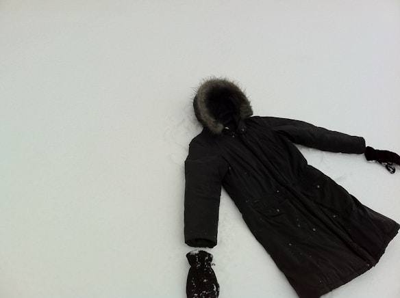 A dark winter coat with fur trim on the hood lies on a flat, snowy surface. The coat appears to have one glove at each sleeve end, suggesting it may have been laid out to mimic a person making a snow angel. The snow is smooth, contrasting with the dark color of the coat.