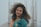 A woman with curly hair wearing a teal and peach traditional outfit, featuring gold embellishments, stands in front of a blurred background that appears to be steel rods or construction materials.
