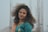 A woman with curly hair wearing a teal and peach traditional outfit, featuring gold embellishments, stands in front of a blurred background that appears to be steel rods or construction materials.