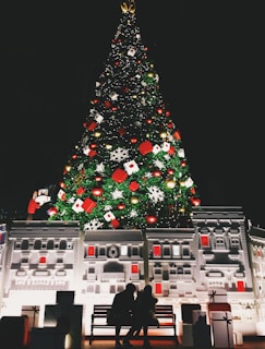 A large, illuminated Christmas tree adorned with various ornaments including red and gold baubles, snowflakes, and gift boxes stands in front of a detailed architectural facade. Two silhouetted figures sit on a bench in front of the display, surrounded by wrapped presents.