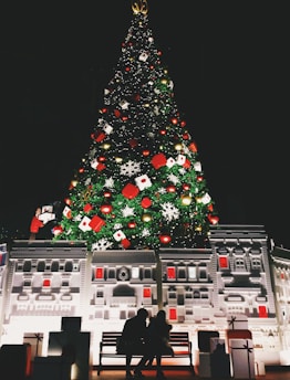 A large, illuminated Christmas tree adorned with various ornaments including red and gold baubles, snowflakes, and gift boxes stands in front of a detailed architectural facade. Two silhouetted figures sit on a bench in front of the display, surrounded by wrapped presents.