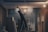 A person on a ladder is working on a ceiling, with a construction or renovation setting in the background. There are exposed light bulbs and various building materials scattered around. The lighting is low, casting shadows, and there is a misty or dusty atmosphere.