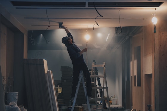 A person on a ladder is working on a ceiling, with a construction or renovation setting in the background. There are exposed light bulbs and various building materials scattered around. The lighting is low, casting shadows, and there is a misty or dusty atmosphere.