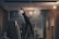 A person on a ladder is working on a ceiling, with a construction or renovation setting in the background. There are exposed light bulbs and various building materials scattered around. The lighting is low, casting shadows, and there is a misty or dusty atmosphere.