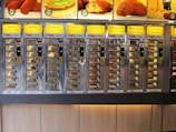 A wall of vending machine compartments offering various fast food items, each with a specific type of snack behind glass doors. The display features bright yellow signs with descriptions of the food items like burgers and kroketten.