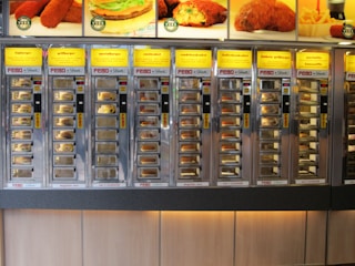 A wall of vending machine compartments offering various fast food items, each with a specific type of snack behind glass doors. The display features bright yellow signs with descriptions of the food items like burgers and kroketten.