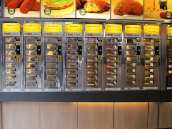 A wall of vending machine compartments offering various fast food items, each with a specific type of snack behind glass doors. The display features bright yellow signs with descriptions of the food items like burgers and kroketten.