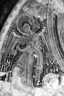A monochromatic fresco depicting an angelic figure with stylized wings and a halo. The figure wears flowing robes and stands against a textured background adorned with script-like symbols. The art has a medieval or early Christian style with intricate details and faded portions that suggest historical aging.