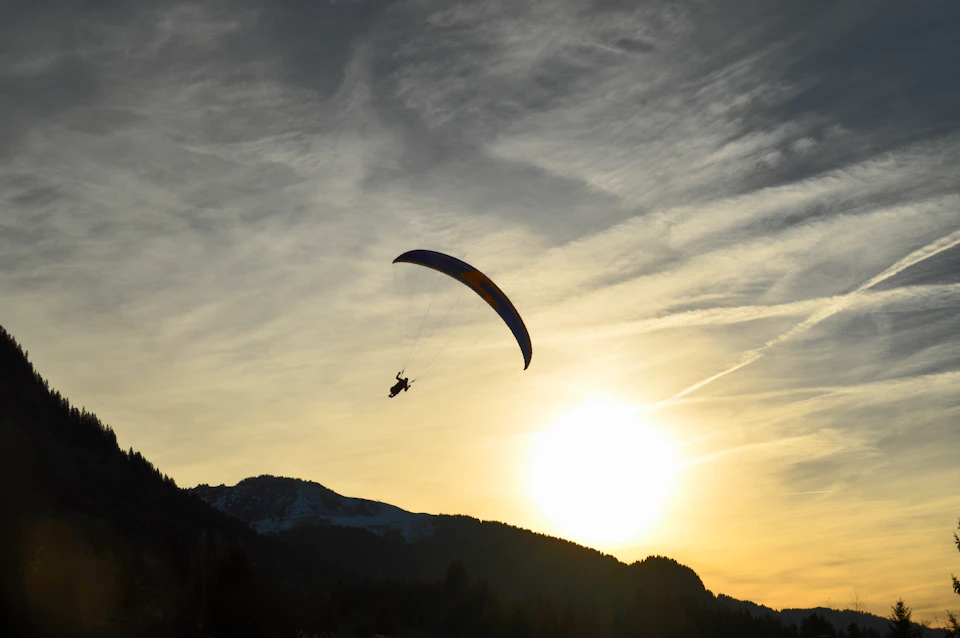 The image depicts a person paragliding over a mountain range at sunset. The paraglider is suspended in mid-air, with their body angled towards the right side of the frame and their feet pointed upwards. Below them, the mountains are bathed in warm hues from the setting sun, creating a stunning backdrop for this airborne adventure. The sky above mirrors the mountain's silhouette, painted in shades of orange and yellow as the sun descends behind the peaks.

The paraglider is positioned centrally within the frame, with their body angled towards the right side to emphasize the vastness of the landscape below them. The mountains stretch out into the distance, their peaks touching the sky at various heights, creating a sense of depth and scale in this breathtaking scene.