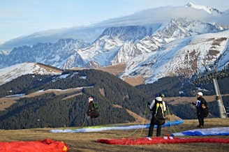 Three individuals prepare for paragliding on a grassy slope with colorful parachutes laid out. Behind them, majestic snow-capped mountains rise against a partly cloudy sky, creating a stunning backdrop. A ski lift tower is visible on the right.