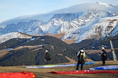 Three individuals prepare for paragliding on a grassy slope with colorful parachutes laid out. Behind them, majestic snow-capped mountains rise against a partly cloudy sky, creating a stunning backdrop. A ski lift tower is visible on the right.