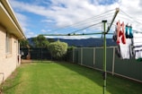 A backyard with lush green grass and a green rotary clothesline holding laundry, including brightly colored socks. The area is enclosed by a green metal fence, and there are shrubs and trees lining the perimeter. A house with beige bricks is visible on the left side, with mountains in the distance under a partly cloudy sky.