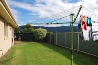 A backyard with lush green grass and a green rotary clothesline holding laundry, including brightly colored socks. The area is enclosed by a green metal fence, and there are shrubs and trees lining the perimeter. A house with beige bricks is visible on the left side, with mountains in the distance under a partly cloudy sky.