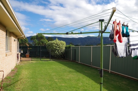 A backyard with lush green grass and a green rotary clothesline holding laundry, including brightly colored socks. The area is enclosed by a green metal fence, and there are shrubs and trees lining the perimeter. A house with beige bricks is visible on the left side, with mountains in the distance under a partly cloudy sky.