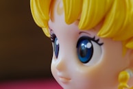 A close-up view of a stylized character figure with large, glossy blue eyes. The figure has yellow hair styled in distinct waves and a smooth complexion typical of anime or cartoon characters.
