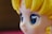 A close-up view of a stylized character figure with large, glossy blue eyes. The figure has yellow hair styled in distinct waves and a smooth complexion typical of anime or cartoon characters.