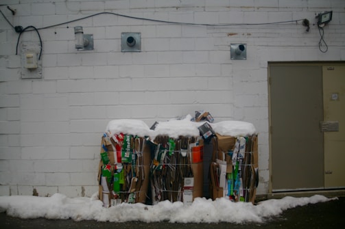 Two large bundles of compressed cardboard and paper are placed against a white brick wall. The top of the bundles is covered in snow, indicating a cold environment. Various pieces of packaging with colorful branding, such as green and red, are visible among the stacks. Electrical conduits and vents are mounted on the wall above the bundles, and to the right, a closed metal door is visible.