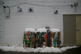 Two large bundles of compressed cardboard and paper are placed against a white brick wall. The top of the bundles is covered in snow, indicating a cold environment. Various pieces of packaging with colorful branding, such as green and red, are visible among the stacks. Electrical conduits and vents are mounted on the wall above the bundles, and to the right, a closed metal door is visible.