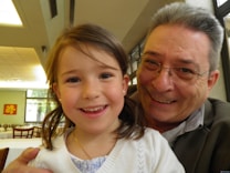 A young girl with brown hair is smiling brightly while an older man with glasses and gray hair is smiling beside her. They appear to be in an indoor dining space with tables and chairs in the background.