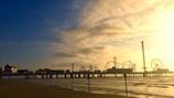 An amusement park extends over a body of water at sunset. The silhouette of roller coasters, rides, and a Ferris wheel is visible against a sky with clouds tinged in golden hues. The ocean waves gently lap at the shoreline, and the pier connecting the park to the beach is clearly delineated.