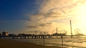 An amusement park extends over a body of water at sunset. The silhouette of roller coasters, rides, and a Ferris wheel is visible against a sky with clouds tinged in golden hues. The ocean waves gently lap at the shoreline, and the pier connecting the park to the beach is clearly delineated.
