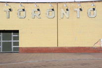 A brick wall with faded yellow paint features the large, block letters spelling out 'TORONTO'. Above the letters are a series of industrial lights. Several patches of paint appear chipped or weathered, and a small section of street pavement is visible in front of the wall.