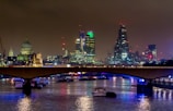 A nighttime cityscape of London is lit up with city lights reflecting off the water. The skyline includes distinctive skyscrapers and historic buildings, and a bridge spans the river, with boats visible beneath.