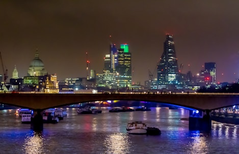 A nighttime cityscape of London is lit up with city lights reflecting off the water. The skyline includes distinctive skyscrapers and historic buildings, and a bridge spans the river, with boats visible beneath.