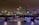 A nighttime cityscape of London is lit up with city lights reflecting off the water. The skyline includes distinctive skyscrapers and historic buildings, and a bridge spans the river, with boats visible beneath.