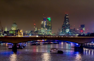 A nighttime shot of London’s business district with illuminated skyscrapers reflecting in the Thames.