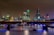A nighttime cityscape of London is lit up with city lights reflecting off the water. The skyline includes distinctive skyscrapers and historic buildings, and a bridge spans the river, with boats visible beneath.