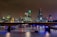 A nighttime cityscape of London is lit up with city lights reflecting off the water. The skyline includes distinctive skyscrapers and historic buildings, and a bridge spans the river, with boats visible beneath.