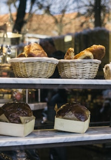 A bakery display window showcases two baskets filled with freshly baked bread at the top shelf and two loaves of dark, round bread in rectangular paper trays on the middle shelf. The warm tones of the bread contrast with the cool reflections on the glass.