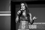 A woman with long hair holds a microphone while standing on a stage. She is wearing a camouflage sports bra, and her toned abs are visible. Her expression appears confident and engaging.