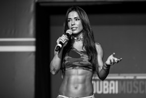 A woman with long hair holds a microphone while standing on a stage. She is wearing a camouflage sports bra, and her toned abs are visible. Her expression appears confident and engaging.
