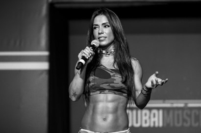 A woman with long hair holds a microphone while standing on a stage. She is wearing a camouflage sports bra, and her toned abs are visible. Her expression appears confident and engaging.