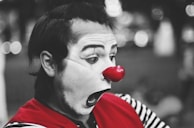 A clown with white face paint and exaggerated facial features is wearing a bright red nose and a red garment over a black and white striped shirt. The focus is on the clown's expressive face, captured in profile with a background of blurred lights.